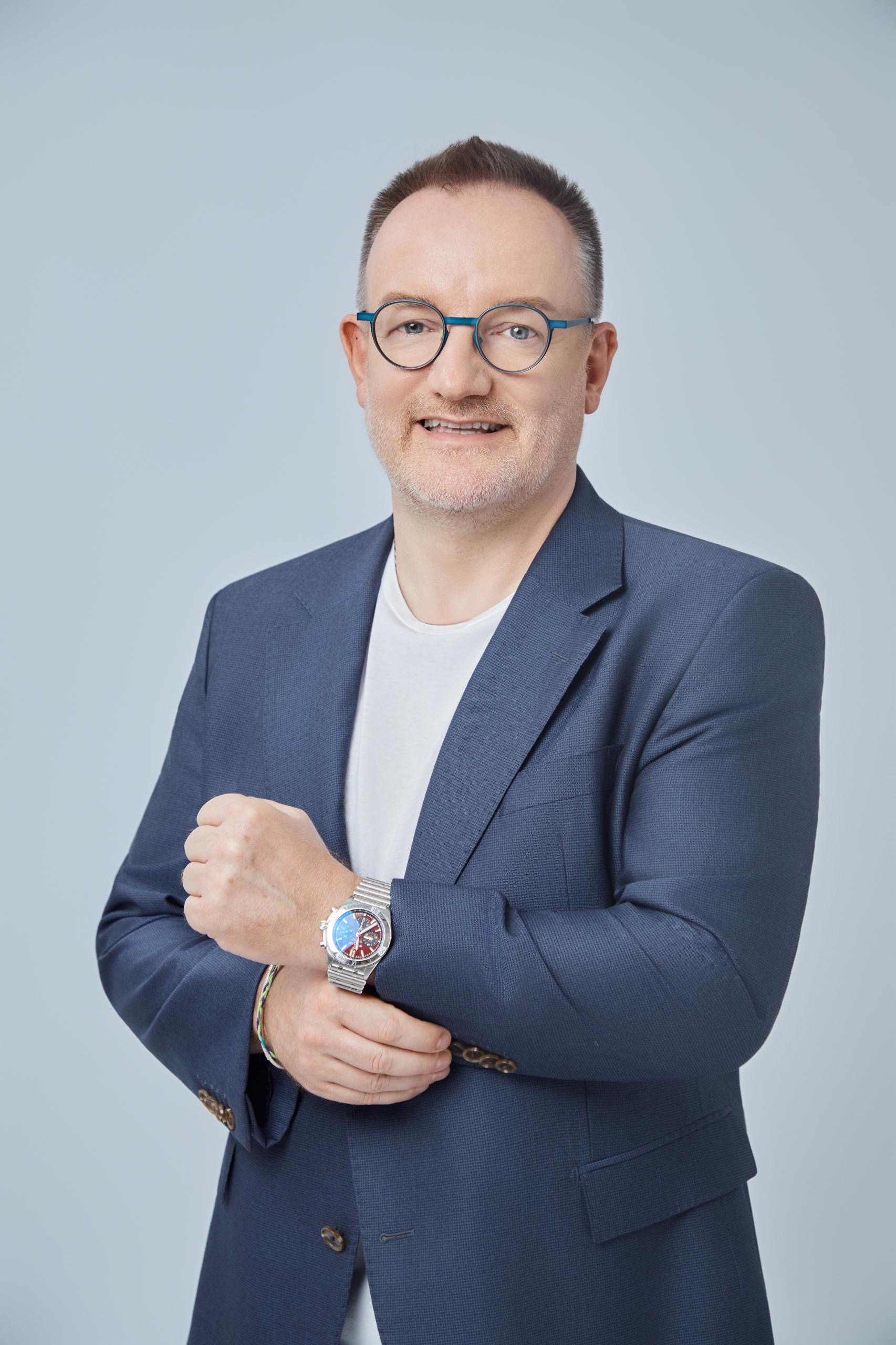 Ein lächelnder Mann mit blauer Brille trägt blauen Anzug und zeigt seine Uhr
